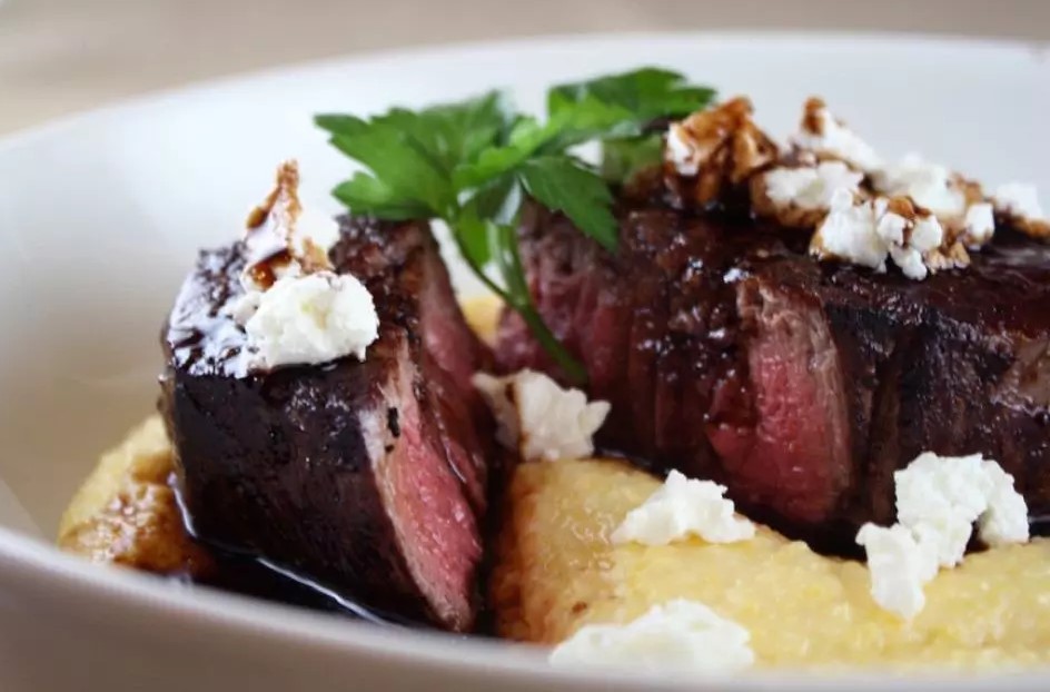 Two thick-cut beef medallions seared to a medium-rare finish, served over a smooth bed of creamy polenta. The dish is topped with crumbled cheese, a drizzle of balsamic glaze, and a sprig of parsley for garnish.
