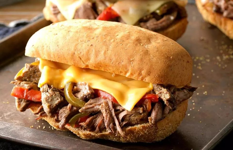 Close-up of a shredded beef sandwich with melted cheese, red and green peppers, served on a toasted roll.