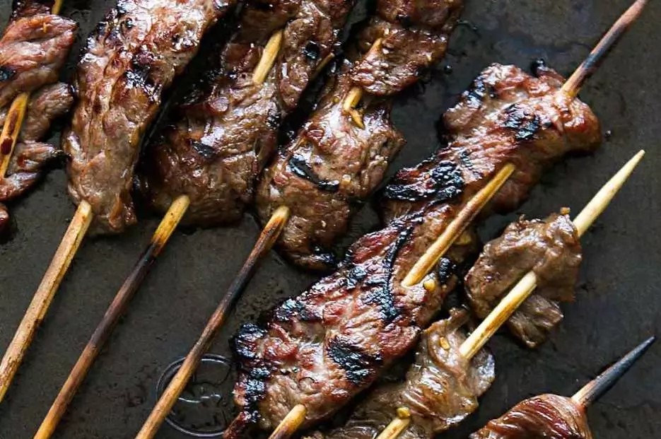 Close-up of several grilled beef skewers, seared with dark char marks and glistening with glaze on wooden sticks.